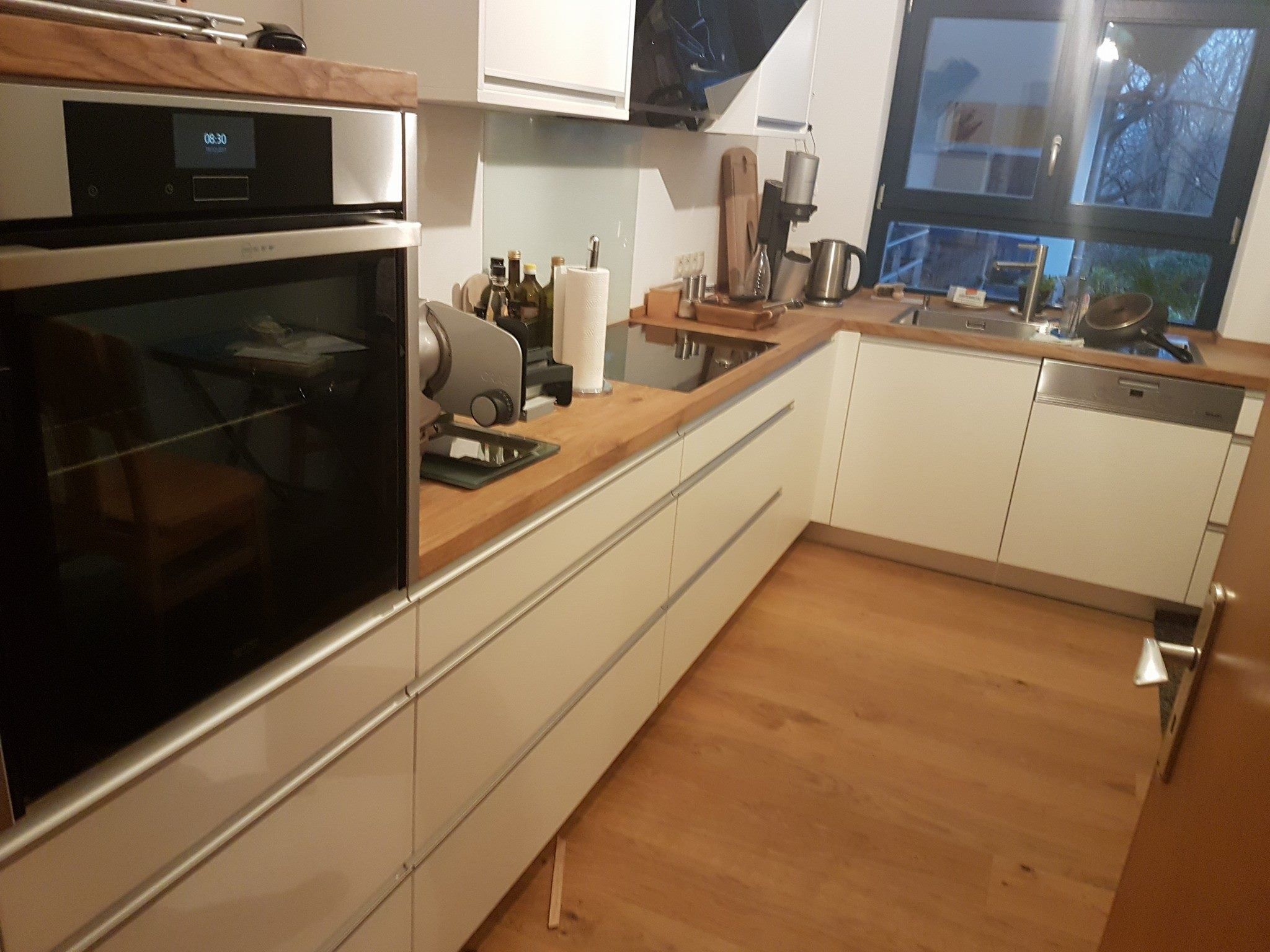 Weiße Küche mit Backofen, Griffleisten und Holzarbeitsplatte, im Hintergrund ein Fenster